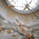 Délicates fleurs artificielles aux pétales blancs et orange sur de fines branches, placées sur un plafond en dôme orné et flou du musée de la Toile de Jouy, surmonté d'un vitrail rond. - Musée de la Toile de Jouy