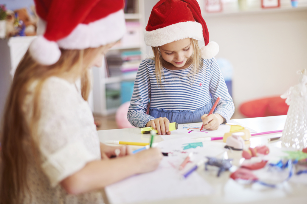 Deux jeunes filles portant des bonnets de Père Noël sont assises à une table, dessinant et coloriant avec des marqueurs. Entourées de matériel artistique, elles s'adonnent à une activité artisanale festive inspirée de l'esprit créatif du musée de la Toile de Jouy. - Musée de la Toile de Jouy