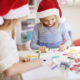 Deux jeunes filles portant des bonnets de Père Noël sont assises à une table, dessinant et coloriant avec des marqueurs. Entourées de matériel artistique, elles s'adonnent à une activité artisanale festive inspirée de l'esprit créatif du musée de la Toile de Jouy. - Musée de la Toile de Jouy