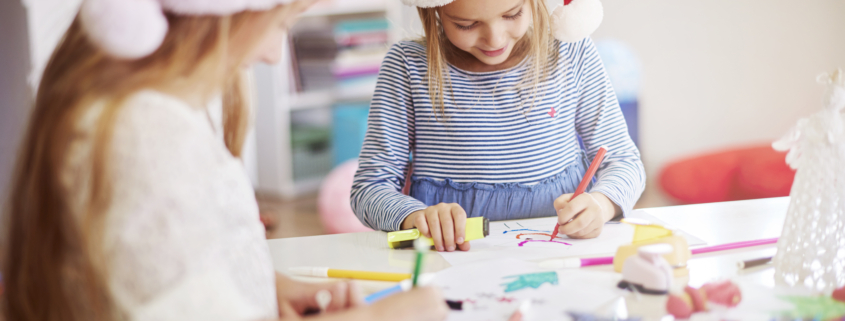 Deux jeunes filles portant des bonnets de Père Noël sont assises à une table, dessinant et coloriant avec des marqueurs. Entourées de matériel artistique, elles s'adonnent à une activité artisanale festive inspirée de l'esprit créatif du musée de la Toile de Jouy. - Musée de la Toile de Jouy