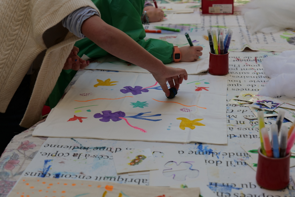 Au musée de la Toile de Jouy, des enfants décorent des tissus à l'aide de marqueurs et de tampons colorés, sur une table recouverte de fournitures artistiques et d'une nappe imprimée. Un enfant presse un tampon sur le tissu, créant ainsi des motifs floraux colorés. - Musée de la Toile de Jouy