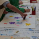 Au musée de la Toile de Jouy, des enfants décorent des tissus à l'aide de marqueurs et de tampons colorés, sur une table recouverte de fournitures artistiques et d'une nappe imprimée. Un enfant presse un tampon sur le tissu, créant ainsi des motifs floraux colorés. - Musée de la Toile de Jouy