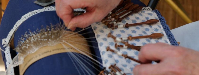 Au musée de la Toile de Jouy, une personne fait de la dentelle avec des fuseaux sur un coussin bleu, en tissant soigneusement des fils. Les mains sont visibles, manipulant habilement les fuseaux en bois pour créer des motifs de dentelle complexes. - Musée de la Toile de Jouy