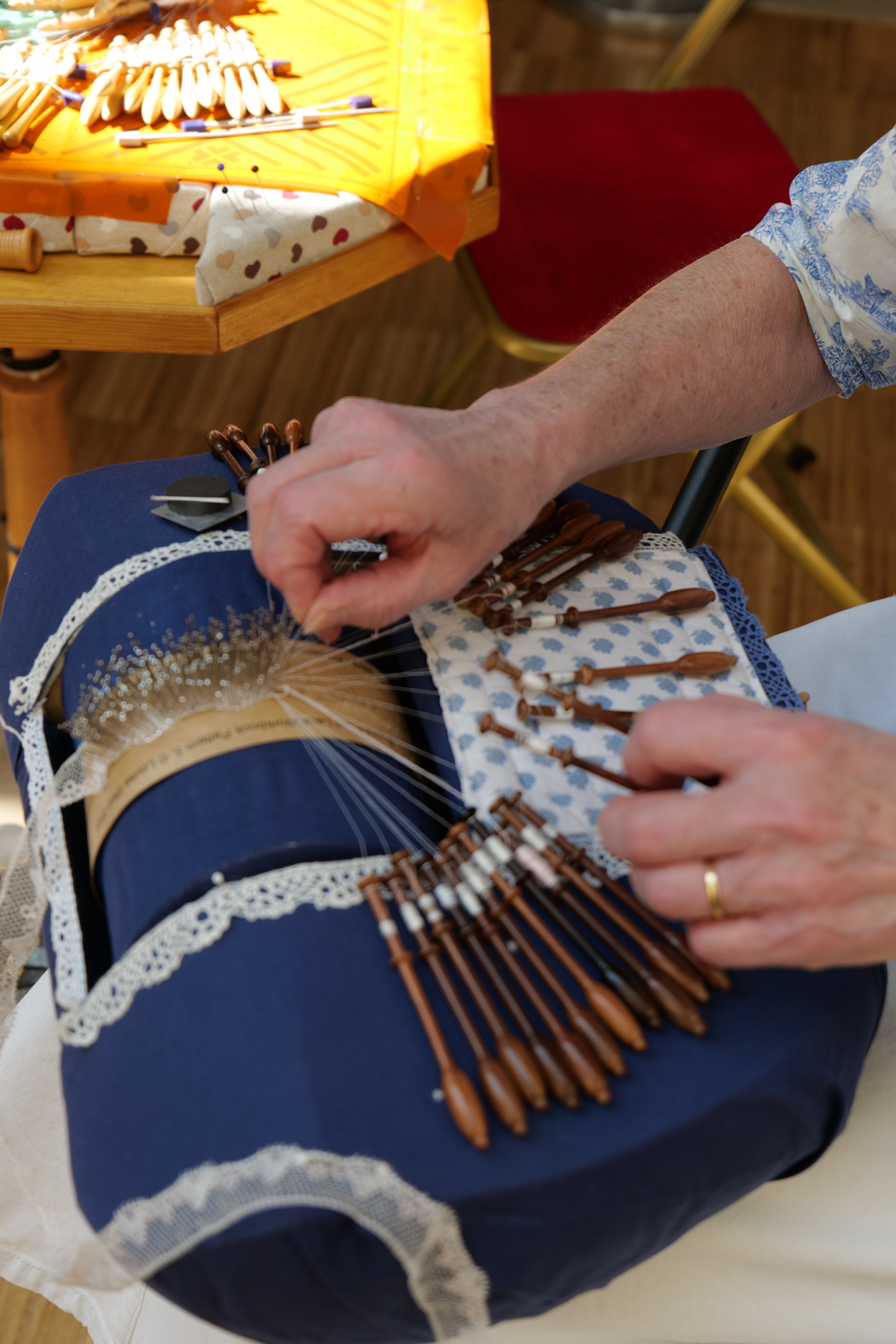 Au musée de la Toile de Jouy, une personne fait de la dentelle avec des fuseaux sur un coussin bleu, en tissant soigneusement des fils. Les mains sont visibles, manipulant habilement les fuseaux en bois pour créer des motifs de dentelle complexes. - Musée de la Toile de Jouy