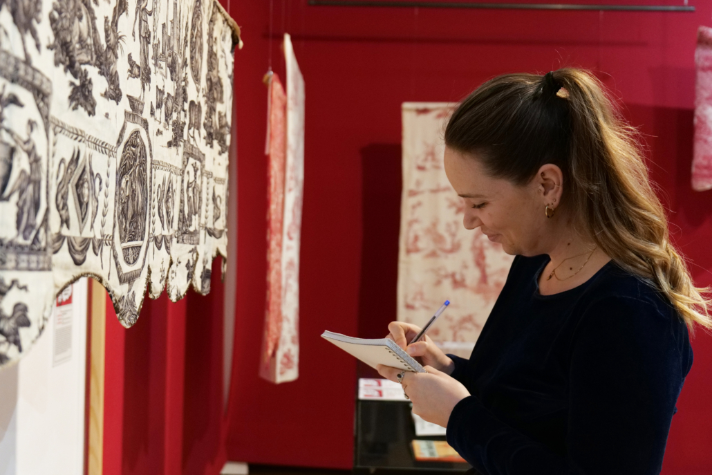 Une femme avec une queue de cheval sourit tout en prenant des notes sur un bloc-notes devant un textile à motifs complexes Made in Jouy exposé sur un mur rouge dans un musée ou une galerie. - Musée de la Toile de Jouy