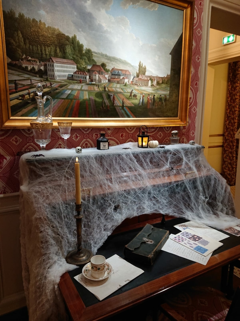 Un piano recouvert de fausses toiles d'araignée est posé sous une grande peinture représentant un champ et un village colorés. Décoré pour Halloween, il contient des bougies, un service à thé et des papiers, créant ainsi une scène vintage et légèrement effrayante, parfaite pour une visite contée. - Musée de la Toile de Jouy