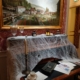 Un piano recouvert de fausses toiles d'araignée est posé sous une grande peinture représentant un champ et un village colorés. Décoré pour Halloween, il contient des bougies, un service à thé et des papiers, créant ainsi une scène vintage et légèrement effrayante, parfaite pour une visite contée. - Musée de la Toile de Jouy