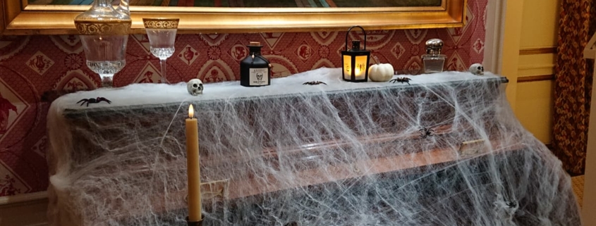 Un piano recouvert de fausses toiles d'araignée est posé sous une grande peinture représentant un champ et un village colorés. Décoré pour Halloween, il contient des bougies, un service à thé et des papiers, créant ainsi une scène vintage et légèrement effrayante, parfaite pour une visite contée. - Musée de la Toile de Jouy