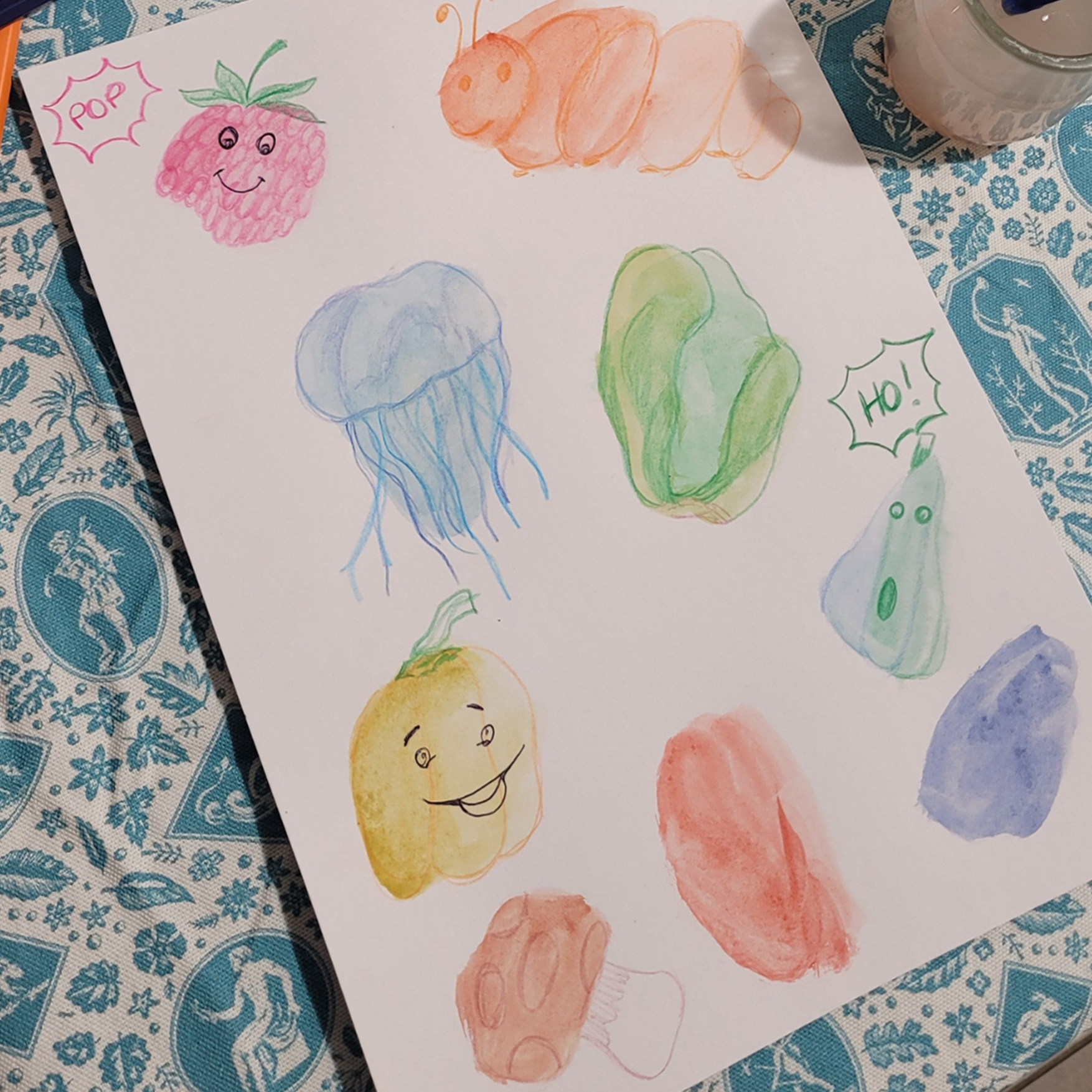 Une feuille de papier posée sur une nappe bleue à motifs de l'Atelier Hirameki présente des gribouillis colorés à l'aquarelle - framboise, chenille, méduse, aubergine et bulles de texte disant "Pop !" et "Ho !" - vous incitant à dessiner tu vois !. - Musée de la Toile de Jouy