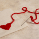 Une aiguille munie d'un fil rouge est enfilée dans un tissu beige, formant un cœur avec le fil. Le fil libre s'étend en une ligne ondulée sur la surface texturée, rappelant le délicat travail manuel du Musée de la Toile de Jouy. - Musée de la Toile de Jouy