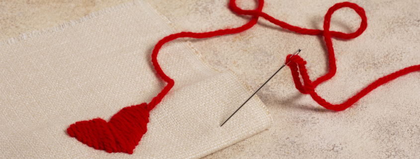 Une aiguille munie d'un fil rouge est enfilée dans un tissu beige, formant un cœur avec le fil. Le fil libre s'étend en une ligne ondulée sur la surface texturée, rappelant le délicat travail manuel du Musée de la Toile de Jouy. - Musée de la Toile de Jouy