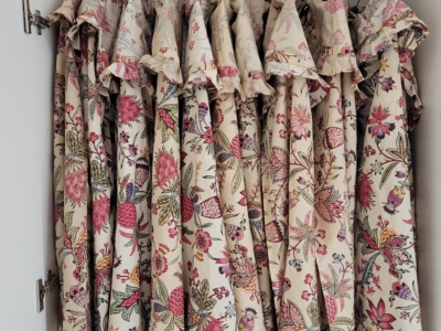 Plusieurs panneaux de rideaux à motifs floraux et à dessus volantés, rappelant les motifs du musée de la Toile de Jouy, sont suspendus étroitement à une tringle métallique à l'intérieur d'une armoire blanche. Le tissu présente des fleurs roses, rouges et vertes sur fond crème. - Musée de la Toile de Jouy