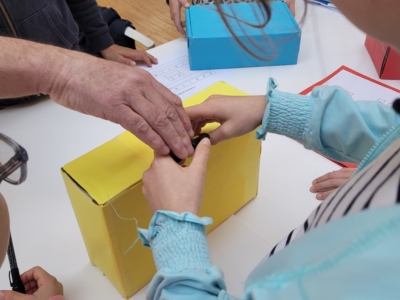 Des enfants et la main d'un adulte interagissent avec des boîtes en carton colorées sur une table du musée de la Toile de Jouy, avec des papiers et des crayons à proximité, ce qui suggère une activité d'apprentissage ou d'artisanat. - Musée de la Toile de Jouy