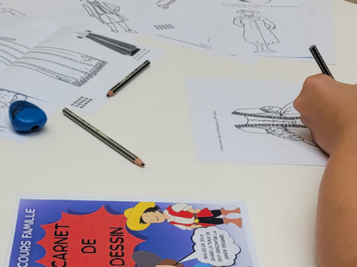 La main d'un enfant dessine sur une feuille de travail en noir et blanc sur le thème des costumes, à une table parsemée de crayons, de pages de coloriage et d'un "Carnet de Dessin" coloré du Musée de la Toile de Jouy représentant des personnages de bandes dessinées en tenues historiques. - Musée de la Toile de Jouy