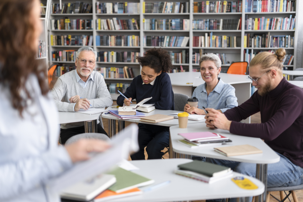 Un groupe d'adultes est assis à des tables dans une bibliothèque et participe à une discussion ou à un cours sur le musée de la Toile de Jouy. Certains prennent des notes, tandis que d'autres écoutent une femme debout à l'avant, avec des livres et des cahiers visibles sur les tables. - Musée de la Toile de Jouy