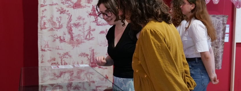 Quatre femmes regardent des documents historiques et des textiles exposés dans une vitrine au Musée de la Toile de Jouy, avec des murs rouges et des panneaux de tissus à motifs en arrière-plan. - Musée de la Toile de Jouy