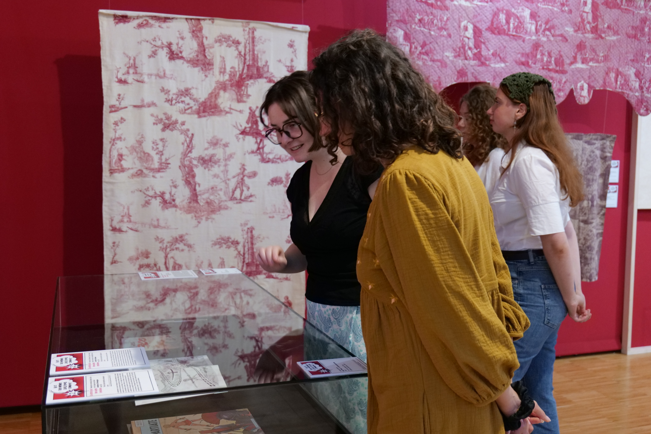 Quatre femmes regardent des documents historiques et des textiles exposés dans une vitrine au Musée de la Toile de Jouy, avec des murs rouges et des panneaux de tissus à motifs en arrière-plan. - Musée de la Toile de Jouy