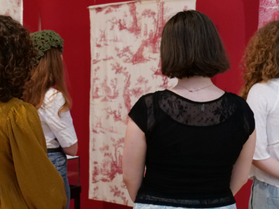 Quatre femmes, dos à la caméra, observent des œuvres d'art textiles à motifs exposées sur des murs rouges dans une galerie du musée de la Toile de Jouy. - Musée de la Toile de Jouy