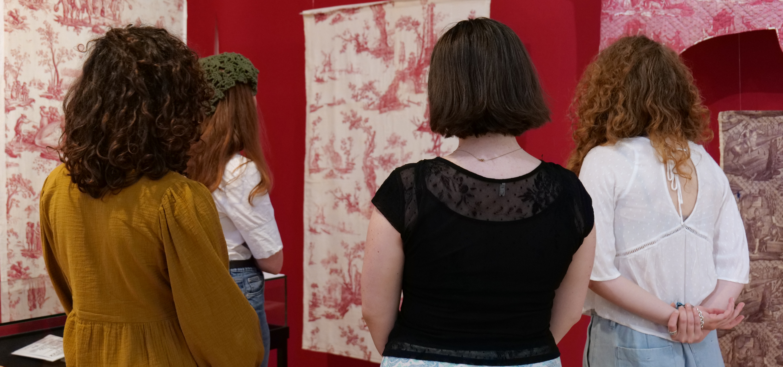 Quatre femmes, dos à la caméra, observent des œuvres d'art textiles à motifs exposées sur des murs rouges dans une galerie du musée de la Toile de Jouy. - Musée de la Toile de Jouy