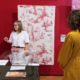 Une femme fait visiter à trois visiteurs la galerie du musée de la Toile de Jouy, où des textiles à motifs rouges et blancs sont exposés sur les murs et derrière des vitres. Les visiteurs écoutent attentivement la guide. - Musée de la Toile de Jouy