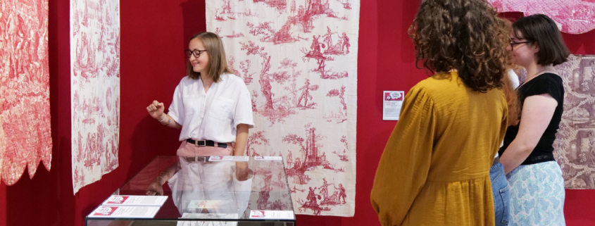 Une femme fait visiter à trois visiteurs la galerie du musée de la Toile de Jouy, où des textiles à motifs rouges et blancs sont exposés sur les murs et derrière des vitres. Les visiteurs écoutent attentivement la guide. - Musée de la Toile de Jouy