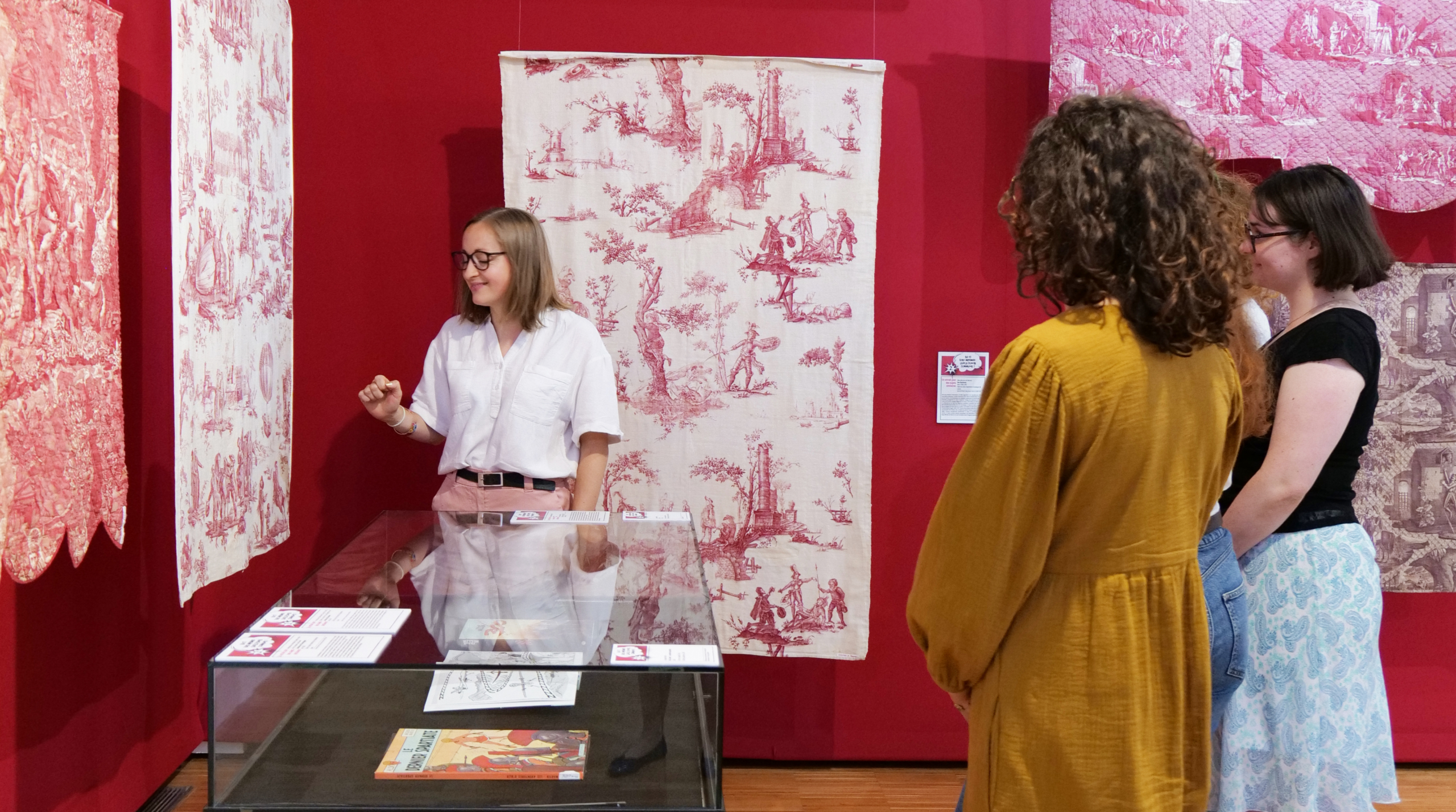 Une femme fait visiter à trois visiteurs la galerie du musée de la Toile de Jouy, où des textiles à motifs rouges et blancs sont exposés sur les murs et derrière des vitres. Les visiteurs écoutent attentivement la guide. - Musée de la Toile de Jouy
