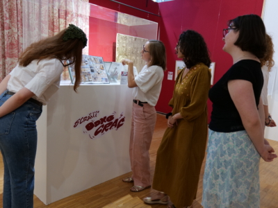 Quatre femmes se trouvent dans une galerie du musée de la Toile de Jouy et regardent une vitrine contenant des œuvres d'art ou des bandes dessinées. Une femme fait un geste vers la vitrine, expliquant quelque chose au groupe. L'exposition est entourée de murs rouges et de rideaux à motifs. - Musée de la Toile de Jouy