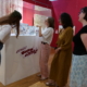 Quatre femmes se trouvent dans une galerie du musée de la Toile de Jouy et regardent une vitrine contenant des œuvres d'art ou des bandes dessinées. Une femme fait un geste vers la vitrine, expliquant quelque chose au groupe. L'exposition est entourée de murs rouges et de rideaux à motifs. - Musée de la Toile de Jouy