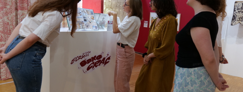 Quatre femmes se trouvent dans une galerie du musée de la Toile de Jouy et regardent une vitrine contenant des œuvres d'art ou des bandes dessinées. Une femme fait un geste vers la vitrine, expliquant quelque chose au groupe. L'exposition est entourée de murs rouges et de rideaux à motifs. - Musée de la Toile de Jouy