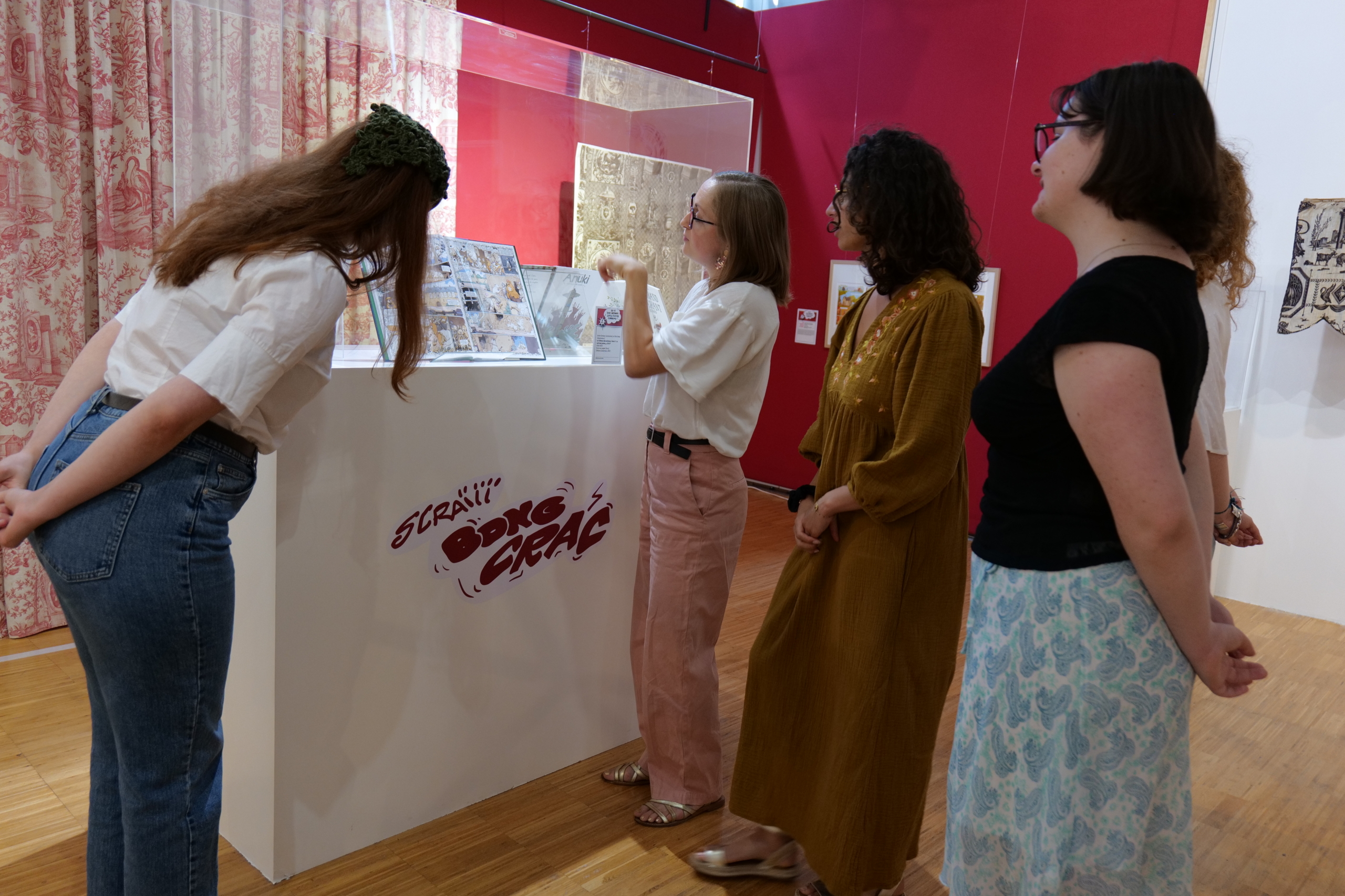 Quatre femmes se trouvent dans une galerie du musée de la Toile de Jouy et regardent une vitrine contenant des œuvres d'art ou des bandes dessinées. Une femme fait un geste vers la vitrine, expliquant quelque chose au groupe. L'exposition est entourée de murs rouges et de rideaux à motifs. - Musée de la Toile de Jouy