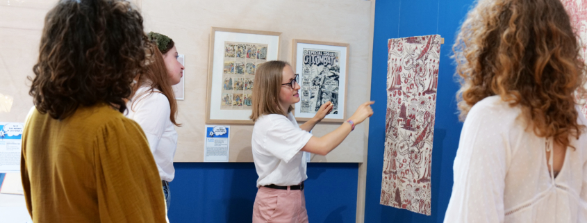 Quatre personnes se trouvent dans la galerie du Musée de la Toile de Jouy, regardant et discutant d'œuvres d'art encadrées et d'un textile à motifs exposé sur des murs bleus. Une personne fait un geste vers le textile, expliquant quelque chose au groupe. - Musée de la Toile de Jouy