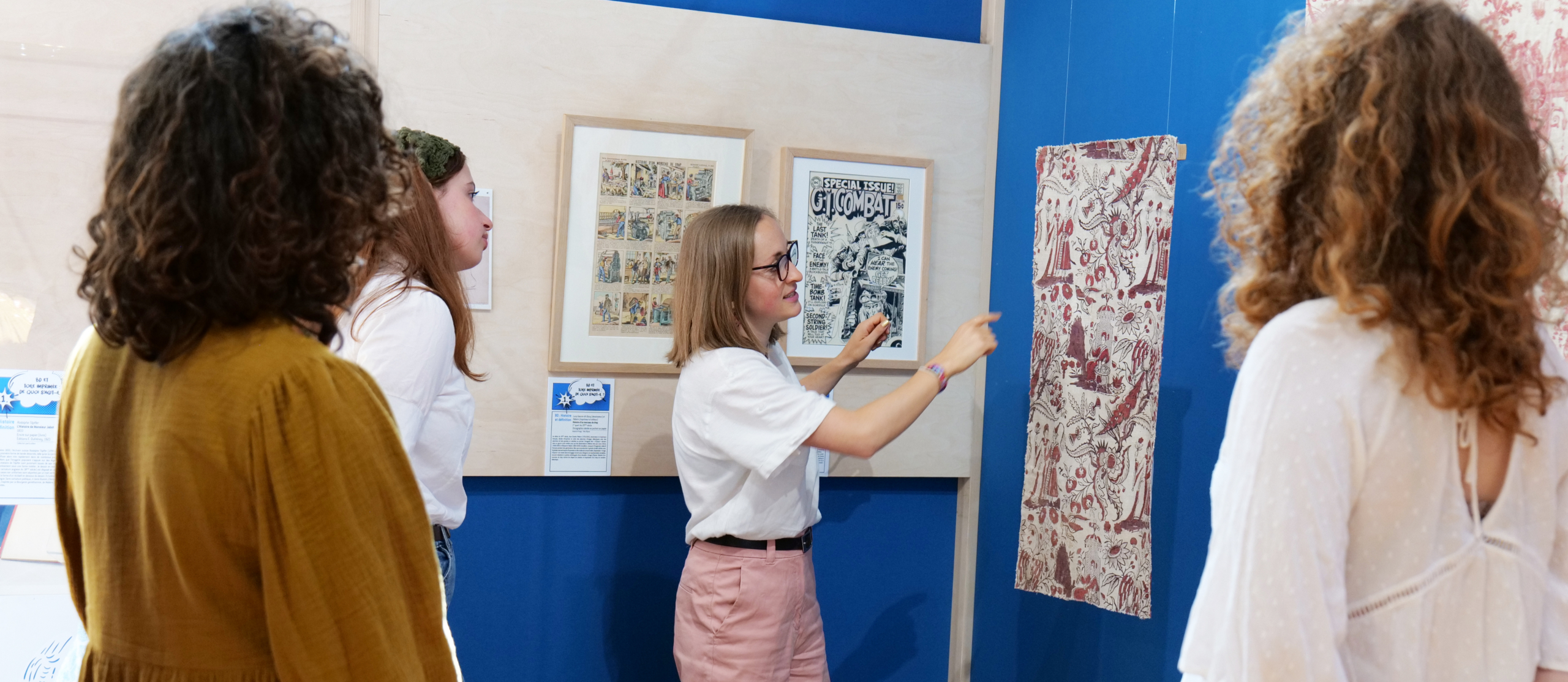 Quatre personnes se trouvent dans la galerie du Musée de la Toile de Jouy, regardant et discutant d'œuvres d'art encadrées et d'un textile à motifs exposé sur des murs bleus. Une personne fait un geste vers le textile, expliquant quelque chose au groupe. - Musée de la Toile de Jouy