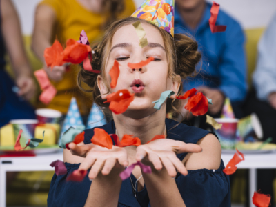 Une jeune fille portant un chapeau de fête souffle des confettis colorés de ses mains lors d'une célébration au Musée de la Toile de Jouy, avec d'autres personnes et des décorations festives visibles à l'arrière-plan. - Musée de la Toile de Jouy