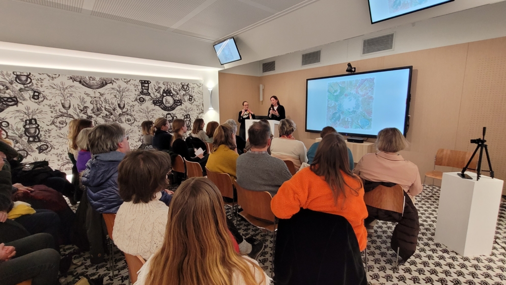 Un groupe de personnes est assis face à deux présentateurs à l'avant d'une salle moderne, parlant et faisant des gestes, tandis que des œuvres d'art colorées du Musée de la Toile de Jouy sont affichées sur un grand écran derrière eux. - Musée de la Toile de Jouy
