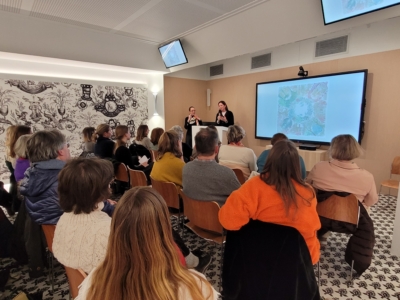 Un groupe de personnes est assis face à deux présentateurs à l'avant d'une salle moderne, parlant et faisant des gestes, tandis que des œuvres d'art colorées du Musée de la Toile de Jouy sont affichées sur un grand écran derrière eux. - Musée de la Toile de Jouy