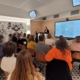 Un groupe de personnes est assis face à deux présentateurs à l'avant d'une salle moderne, parlant et faisant des gestes, tandis que des œuvres d'art colorées du Musée de la Toile de Jouy sont affichées sur un grand écran derrière eux. - Musée de la Toile de Jouy
