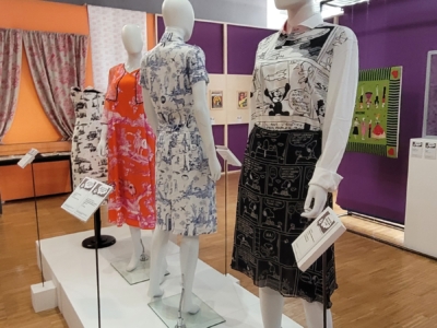 Trois mannequins présentent des robes à motifs dans une exposition du musée de la Toile de Jouy aux murs violets et orange, au sol en bois et aux panneaux d'information placés à côté de chaque mannequin. L'espace est très éclairé. - Musée de la Toile de Jouy