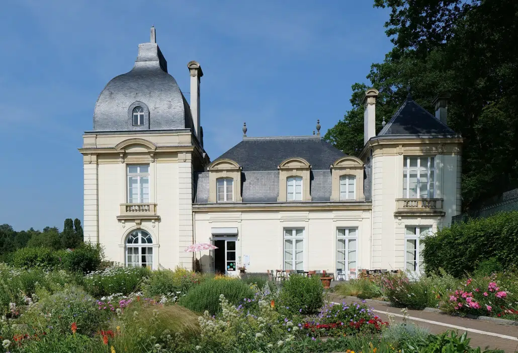 Un élégant manoir de couleur claire avec deux tours et un toit en ardoise, entouré de jardins luxuriants et de fleurs colorées sous un ciel bleu clair. Des tables et des chaises sont installées près de l'entrée. - Musée de la Toile de Jouy