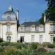 Un élégant manoir de couleur claire avec deux tours et un toit en ardoise, entouré de jardins luxuriants et de fleurs colorées sous un ciel bleu clair. Des tables et des chaises sont installées près de l'entrée. - Musée de la Toile de Jouy