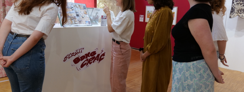 Cinq femmes se tiennent autour d'une vitrine blanche contenant des bandes dessinées. Elles discutent lors d'une visite guidée dans une exposition d'art aux murs rouges et blancs et au sol en bois. - Musée de la Toile de Jouy