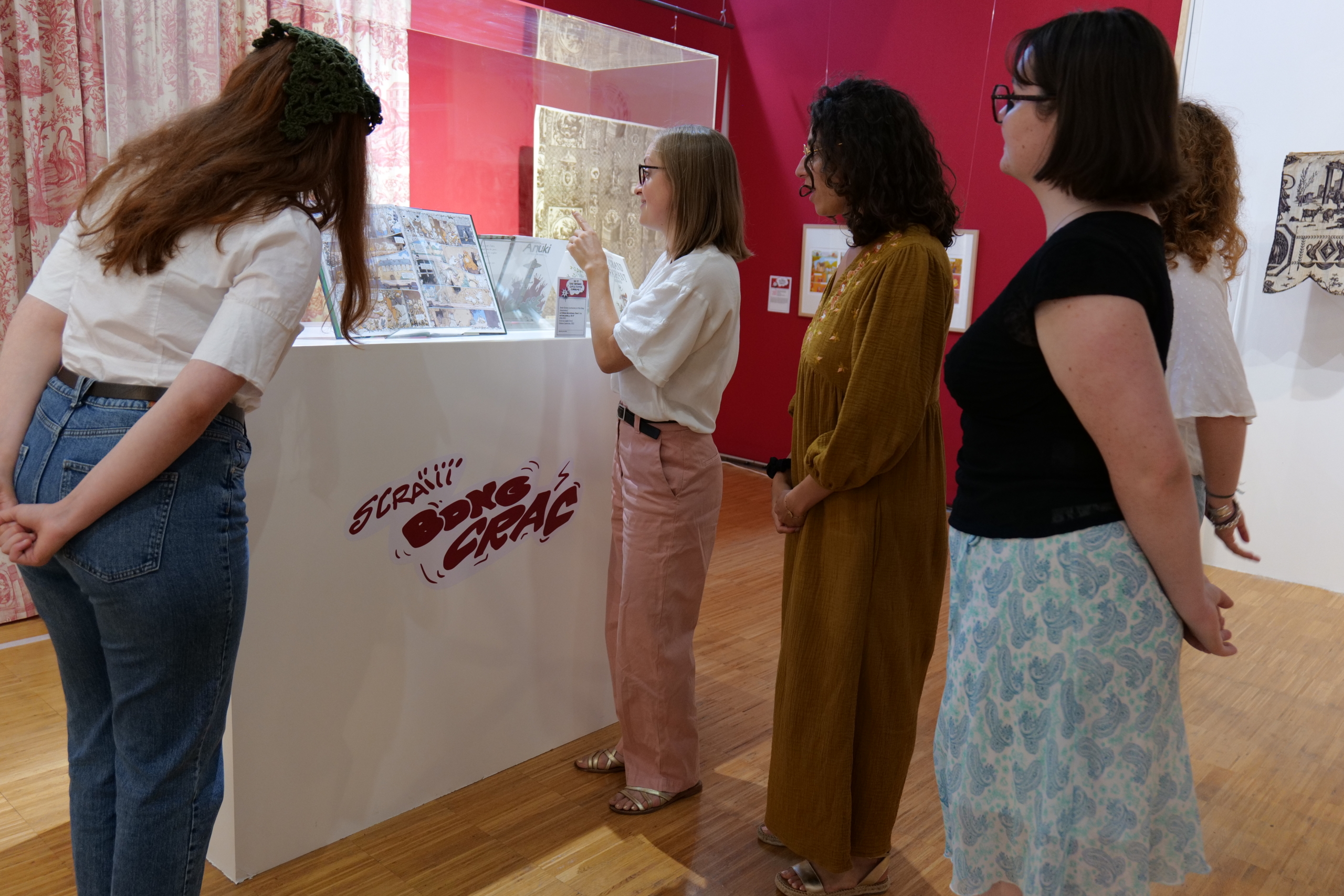 Cinq femmes se tiennent autour d'une vitrine blanche contenant des bandes dessinées. Elles discutent lors d'une visite guidée dans une exposition d'art aux murs rouges et blancs et au sol en bois. - Musée de la Toile de Jouy