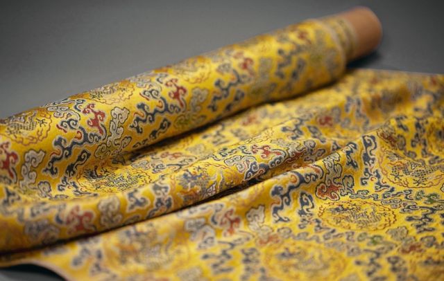 Rouleau de tissu de brocart jaune présentant des motifs complexes de dragons et de nuages en or, bleu et rouge, partiellement déroulé sur une surface plane. - Musée de la Toile de Jouy