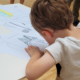 Un jeune enfant aux cheveux bruns courts se penche sur une table, coloriant ou dessinant sur une feuille de dessin imprimée - des activités enfants parfaites pour garder les enfants occupés pendant les vacances scolaires. D'autres feuilles de dessin et les jambes d'une personne sont visibles à l'arrière-plan. - Musée de la Toile de Jouy