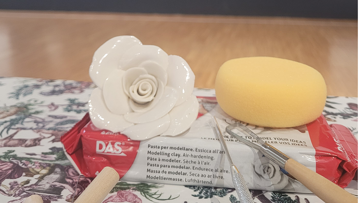 Une rose en argile blanche, fabriquée lors d'un atelier de modelage, est posée sur un bloc rouge de pâte à modeler DAS, avec une éponge jaune, des outils de sculpture et un tapis à motifs floraux à côté, le tout posé sur un sol en bois et un mur uni. - Musée de la Toile de Jouy