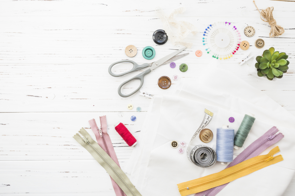 Vue à plat des fournitures de couture de Faty Style Atelier couture sur une surface en bois blanc, comprenant des ciseaux, des fermetures à glissière, des bobines de fil, des boutons, du tissu, un mètre ruban, des aiguilles, du fil rouge, une roue d'épingle et une petite plante succulente en pot. - Musée de la Toile de Jouy