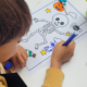 Un enfant colorie au feutre un dessin représentant un squelette sur le thème d'Halloween. Sur la feuille figurent également une citrouille, une araignée, des étoiles et des bonbons. Des crayons de couleur et une gomme se trouvent à proximité, sur la table. - Musée de la Toile de Jouy