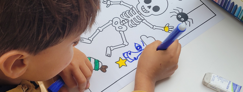 Un enfant colorie au feutre un dessin représentant un squelette sur le thème d'Halloween. Sur la feuille figurent également une citrouille, une araignée, des étoiles et des bonbons. Des crayons de couleur et une gomme se trouvent à proximité, sur la table. - Musée de la Toile de Jouy