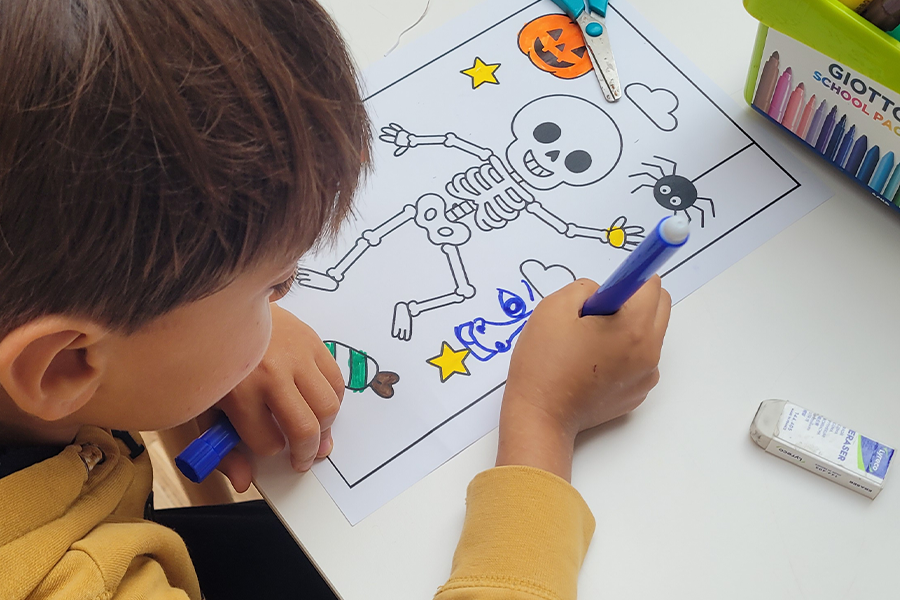 Un enfant colorie au feutre un dessin représentant un squelette sur le thème d'Halloween. Sur la feuille figurent également une citrouille, une araignée, des étoiles et des bonbons. Des crayons de couleur et une gomme se trouvent à proximité, sur la table. - Musée de la Toile de Jouy