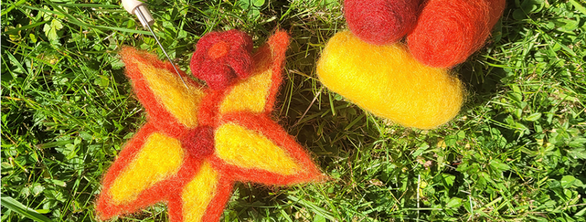 Une étoile en feutre jaune et rouge ainsi que des pelotes de laine sont posées sur de l'herbe verte ; on aperçoit une aiguille à feutrer dans un coin, ce qui évoque l'ambiance d'un atelier pour enfants, idéale pour s'initier à l'art de la laine feutrée. - Musée de la Toile de Jouy
