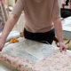 Une personne mesure et dispose des fleurs et du tissu « Toile de Jouy » sur une table, avec des ciseaux à portée de main et d’autres rouleaux de tissu ainsi que du matériel de loisirs créatifs à l’arrière-plan, créant ainsi une charmante ambiance d’atelier pour adultes, éclairée par une petite lampe. - Musée de la Toile de Jouy